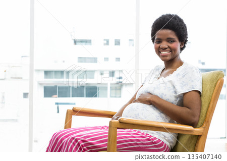 Pregnant African American woman resting hands on abdomen while smiling in wooden armchair by window Pregnant African American woman resting hands on abdomen while smiling in wooden armchair by window 135407140