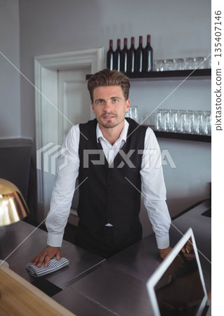 Man wearing white shirt and black vest cleaning stainless steel counter with striped cloth at bar 135407146