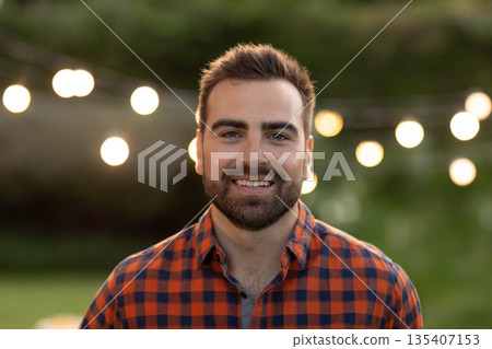 Man standing in backyard garden at dusk wearing checkered shirt under glowing string lights 135407153