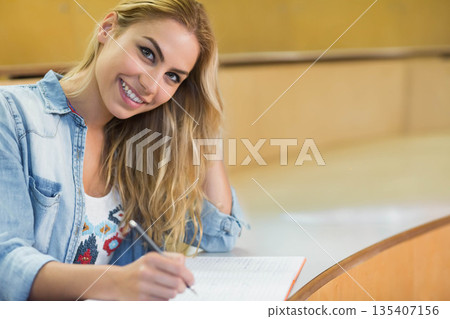 Female student writing in notebook, resting head on hand at curved desk in college lecture hall 135407156