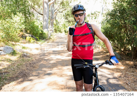 White male cyclist standing on wooded trail holding bike drinking from hydration pack, copy space 135407164