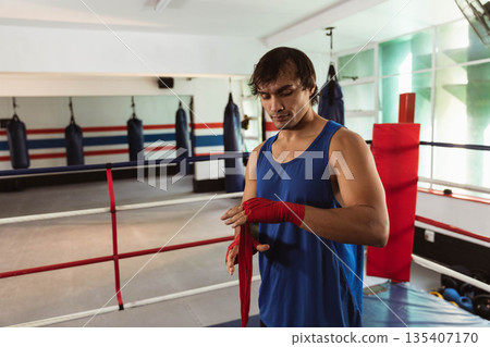 Male boxer wrapping red hand wraps around wrists inside boxing ring with punching bags, copy space Male boxer wrapping red hand wraps around wrists inside boxing ring with punching bags, copy space 135407170