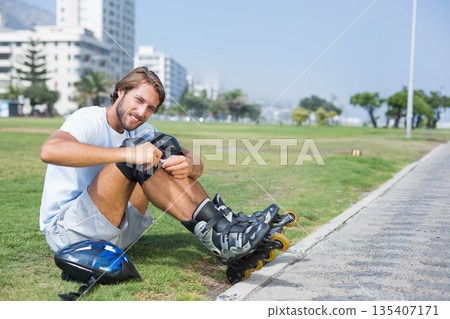 Male fastening knee pad beside park walkway, inline skates, wrist guards, helmet, copy space Male fastening knee pad beside park walkway, inline skates, wrist guards, helmet, copy space 135407171