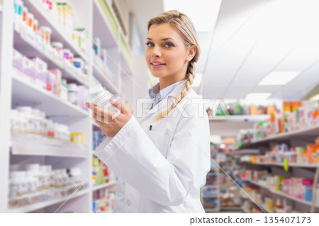 Female pharmacist wearing white lab coat in pharmacy aisle checking pill bottle label Female pharmacist wearing white lab coat in pharmacy aisle checking pill bottle label 135407173