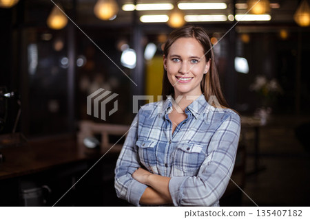 Woman standing with arms crossed in modern co-working space under pendant lamps near wooden desks 135407182