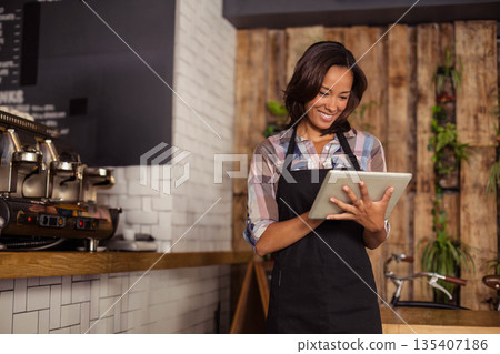 African American Asian barista in apron standing at coffee counter using tablet by espresso machine 135407186