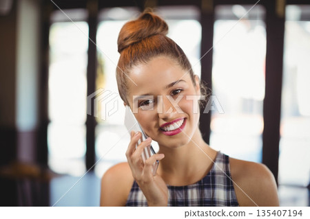 Woman talking on white smartphone while smiling inside cafe lounge near bright glass windows Woman talking on white smartphone while smiling inside cafe lounge near bright glass windows 135407194