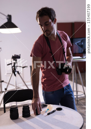 Mid adult man standing in studio beside table organizing DSLR camera and lenses for studio shoot Mid adult man standing in studio beside table organizing DSLR camera and lenses for studio shoot 135407199