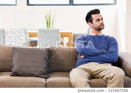 Man sitting on tan sofa at home with potted plant and wooden dining table, copy space 135407208