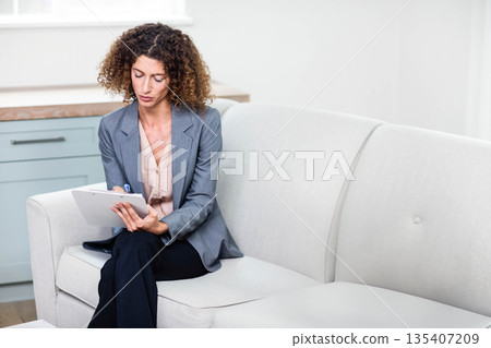 Woman wearing grey blazer sitting on light sofa in office lounge writing on clipboard with pen 135407209