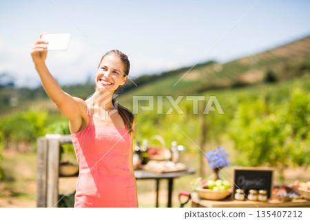 Woman standing at vineyard stand wearing dress taking selfie with smartphone by baskets, copy space 135407212
