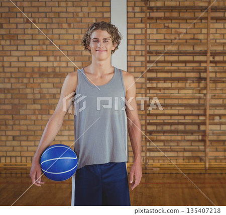 Teenage boy athlete smiling while holding blue basketball against brick wall in school gym Teenage boy athlete smiling while holding blue basketball against brick wall in school gym 135407218