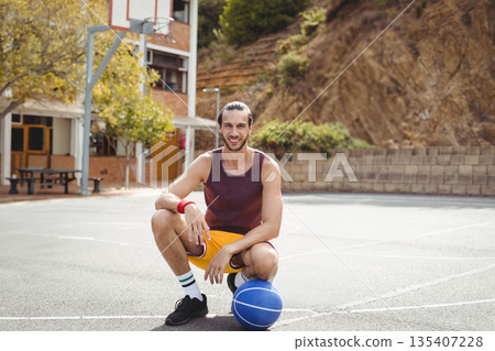 Adult man in sportswear kneeling at outdoor basketball court gripping ball near metal hoop 135407228