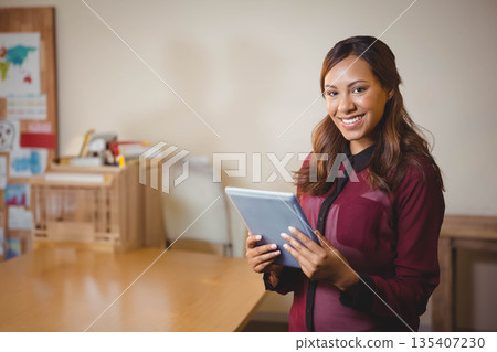 Woman wearing maroon blouse holding tablet standing by wooden table in classroom, copy space Woman wearing maroon blouse holding tablet standing by wooden table in classroom, copy space 135407230