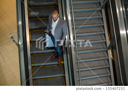 Man standing on down escalator with glass rail at terminal holding suitcase in blazer, copy space 135407231