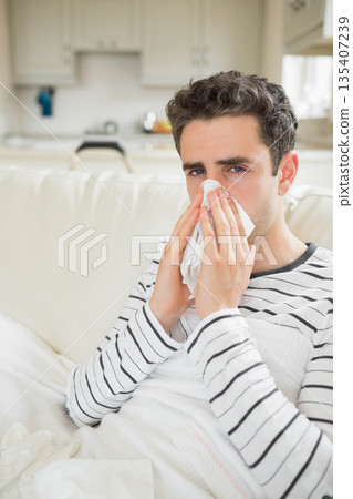Man in early 30s blowing nose into tissue sitting on sofa with white blanket near kitchen Man in early 30s blowing nose into tissue sitting on sofa with white blanket near kitchen 135407239