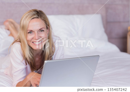 Female lying on stomach on bed in bedroom using laptop smiling at viewer with pillows 135407242