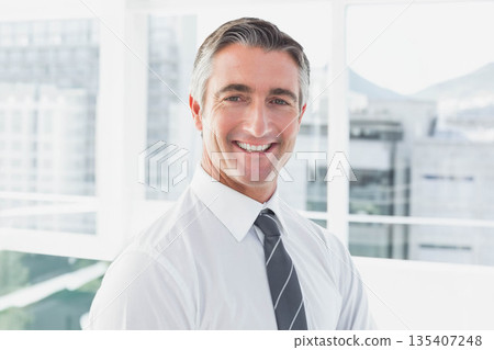 Mature man in white shirt and tie standing in office by floor-to-ceiling windows 135407248