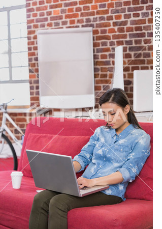 Chinese woman holding coffee cup and typing on laptop near flip chart in loft, copy space 135407250