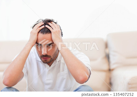 Man leaning forward in living room gripping head by sofa in polo shirt and jeans 135407252