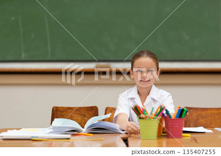 Female child student studying at wooden desk in classroom with open workbook, pencils and pens Female child student studying at wooden desk in classroom with open workbook, pencils and pens 135407255