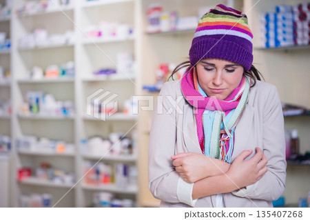 Woman in twenties standing in pharmacy near medicine shelves wearing beanie and scarves, copy space Woman in twenties standing in pharmacy near medicine shelves wearing beanie and scarves, copy space 135407268