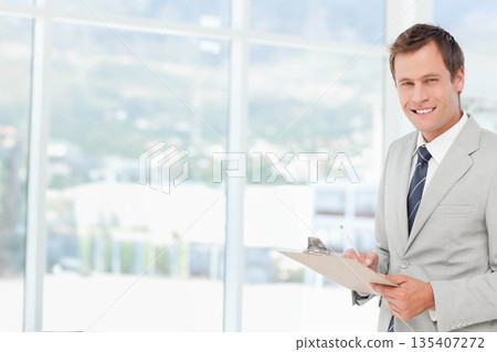 Man wearing suit standing near glass windows in office holding clipboard and pen, copy space Man wearing suit standing near glass windows in office holding clipboard and pen, copy space 135407272