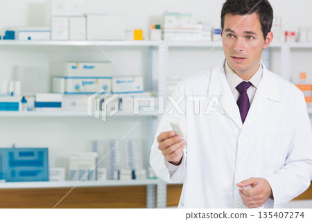 Mature male pharmacist wearing lab coat consulting and holding medicine box in pharmacy, copy space 135407274
