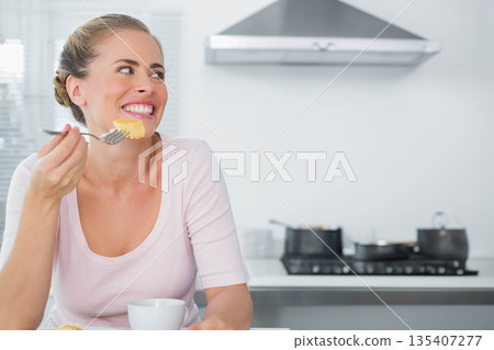 Woman eating food with fork near white mug at kitchen counter, smiling looking right, copy space 135407277