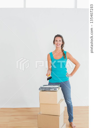 Woman holding paintbrush and painting white wall at home with paint tray on cardboard boxes 135407283