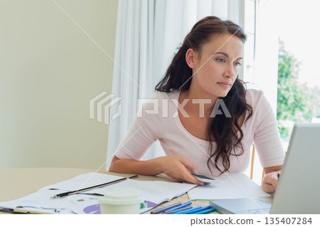 Female remote worker holding payment card while scanning laptop screen at home desk with coffee cup 135407284