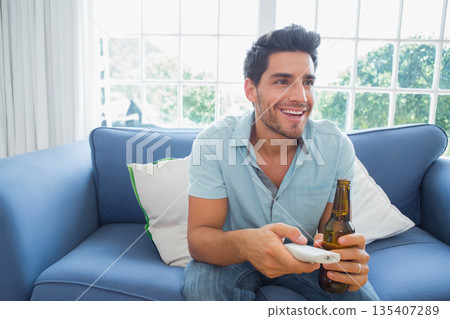 Man holding beer bottle, TV remote control sitting on blue sofa near window showing garden view Man holding beer bottle, TV remote control sitting on blue sofa near window showing garden view 135407289