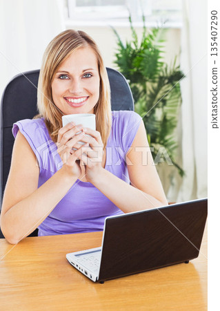 Woman sitting at wooden desk in home office smiling while holding mug near face with laptop 135407290