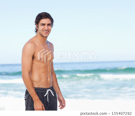 Shirtless man standing on sandy beach at water's edge wearing dark swim trunks amid ocean waves Shirtless man standing on sandy beach at water's edge wearing dark swim trunks amid ocean waves 135407292