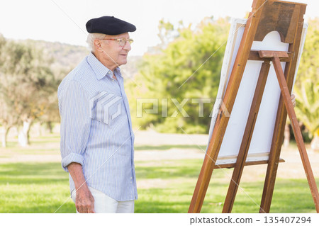 Senior man painter studying blank canvas on easel in grassy park wearing beret, glasses, copy space Senior man painter studying blank canvas on easel in grassy park wearing beret, glasses, copy space 135407294