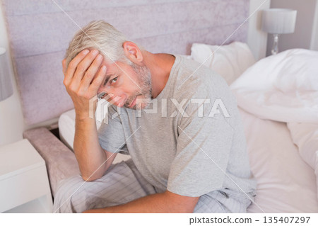 Middle-aged man sitting on bed edge pressing forehead by nightstand lamp wearing gray pajamas 135407297