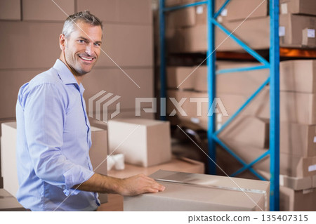 Man in his forties packing cardboard boxes at warehouse table with tape dispenser, copy space 135407315
