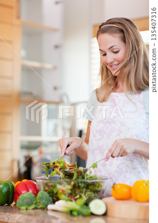 Woman wearing floral apron tossing leafy greens in clear glass bowl on kitchen countertop Woman wearing floral apron tossing leafy greens in clear glass bowl on kitchen countertop 135407316