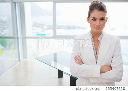 Businesswoman in blazer standing with arms crossed by table and windows in boardroom, copy space 135407318