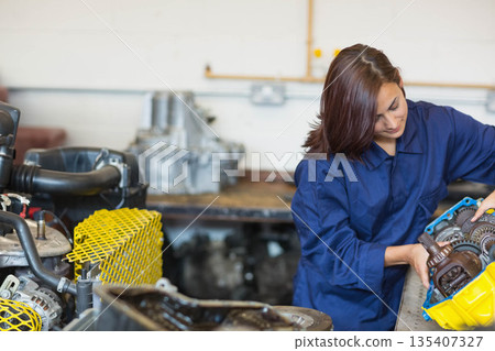 Woman in coverall examining gearbox housing at workbench with gears and safety guard, copy space Woman in coverall examining gearbox housing at workbench with gears and safety guard, copy space 135407327