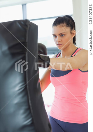 Woman wearing pink sports top navy leggings black boxing gloves punching heavy bag at gym windows Woman wearing pink sports top navy leggings black boxing gloves punching heavy bag at gym windows 135407328