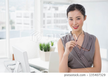 Chinese woman in grey dress standing in office near window holding eyeglasses by desk, copy space Chinese woman in grey dress standing in office near window holding eyeglasses by desk, copy space 135407329