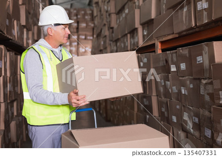 Male warehouse worker lifting cardboard box onto hand truck in warehouse aisle in reflective vest Male warehouse worker lifting cardboard box onto hand truck in warehouse aisle in reflective vest 135407331