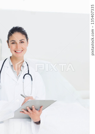 Female doctor standing beside bed with stethoscope holding clipboard and pen in clinic, copy space Female doctor standing beside bed with stethoscope holding clipboard and pen in clinic, copy space 135407335