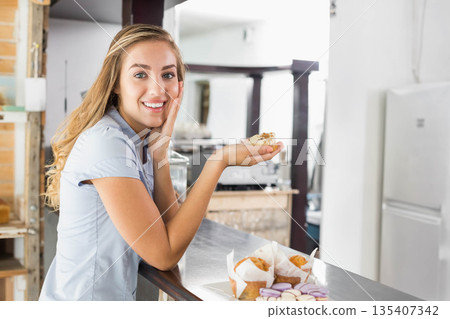 Woman leaning on metal counter in small cafe, holding pastry near plate of muffins and macarons Woman leaning on metal counter in small cafe, holding pastry near plate of muffins and macarons 135407342