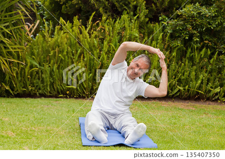 Senior male in sportswear sitting on blue exercise mat on grass lawn in garden stretching muscles Senior male in sportswear sitting on blue exercise mat on grass lawn in garden stretching muscles 135407350