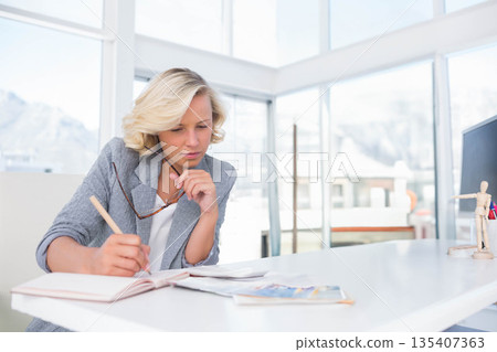 Professional woman wearing gray blazer writing in notebook and holding eyeglasses at office desk Professional woman wearing gray blazer writing in notebook and holding eyeglasses at office desk 135407363