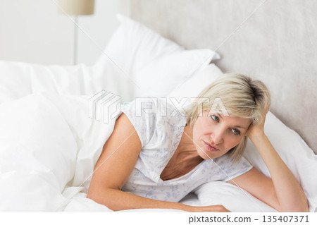 Middle-aged woman reclining on bed in bedroom, leaning back on white duvet under bedside lamp 135407371
