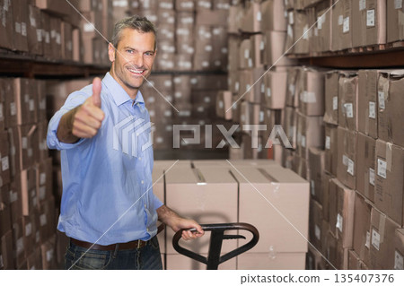 Middle-aged man pushing pallet jack with labeled boxes between metal racks, showing thumbs-up 135407376