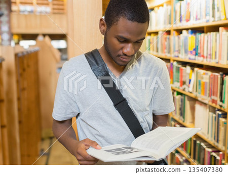 African American male student reading open book in university library carrying black shoulder bag African American male student reading open book in university library carrying black shoulder bag 135407380